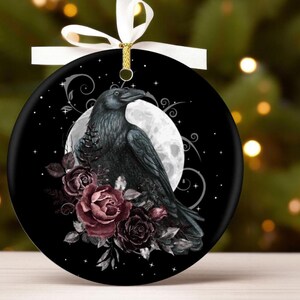 May include: A round black ceramic ornament featuring a detailed illustration of a black raven perched before a large white full moon. The design includes dark red roses and swirling silver filigree accents. A white ribbon is tied at the top.