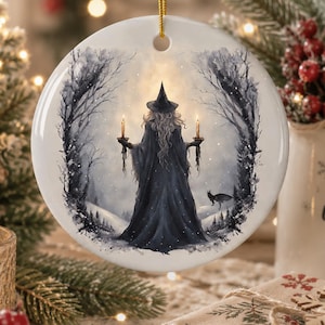Puede incluir: Un adorno de cerámica blanca redonda con una ilustración impresa de una figura con capa oscura y sombrero puntiagudo en un bosque nevado. La figura sostiene dos velas encendidas y se ve un pequeño gato negro cerca. Cuelga de un cordón dorado.