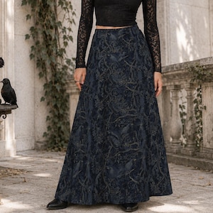May include: A black long-sleeved lace crop top with a V-neckline paired with a floor-length dark blue maxi skirt featuring a detailed tonal pattern of branches and birds. The outfit is completed with a black choker necklace and black shoes.