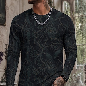 Pode incluir: Uma camisola de manga comprida com gola redonda com um estampado de floresta escura com ramos entrela&ccedil;ados, folhas e silhuetas subtis de p&aacute;ssaros em tons de verde escuro, carv&atilde;o e preto, combinada com cal&ccedil;as de ganga pretas rasgadas e uma corrente prateada.