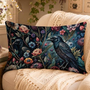 Op de afbeelding: Een rechthoekig sierkussen met een donkerblauwe achtergrond, een levendig bloemenpatroon en twee zwarte raven. Het ontwerp bevat diverse kleurrijke bloemen in tinten roze, paars en oranje, afgewisseld met groen gebladerte.