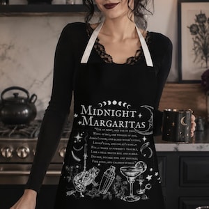 May include: A black apron featuring white graphic illustrations of a frog, a cocktail shaker, and a margarita glass. The text reads Midnight Margaritas along with a poem. The apron has white neck straps and is worn over a black long-sleeve shirt.