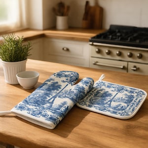 Op de afbeelding: Een blauw-witte ovenwant en een vierkante pannenlap met een toile de jouy-patroon met illustraties van de Eiffeltoren en bloemmotieven. Beide items hebben een witte stoffen rand en een lusje om ze aan op te hangen.