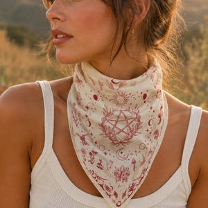 May include: A triangular bandana in off-white fabric featuring a detailed maroon-colored print. The design includes a central pentagram surrounded by celestial motifs, including suns, moons, stars, and various botanical elements like mushrooms.