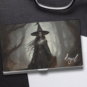May include: A metal business card holder with a dark forest scene featuring a witch in a black dress and pointed hat. The text "Angel" is printed on the card holder.