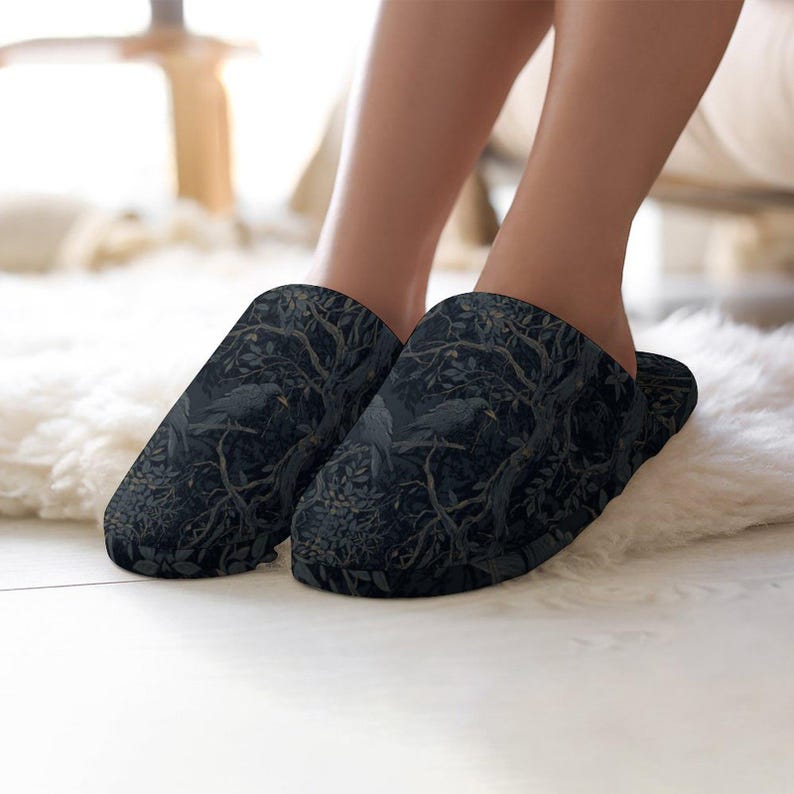 May include: A pair of black slippers with a dark forest and raven design. The slippers are made of a soft material and are designed for indoor use. The slippers are shown on a white fluffy rug.