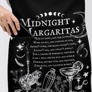 May include: A black apron featuring white graphic illustrations of a moon, stars, a frog, a cocktail shaker, and a margarita glass. The text reads Midnight Margaritas followed by a poem starting with Eye of newt, and toe of frog, wool of bat.