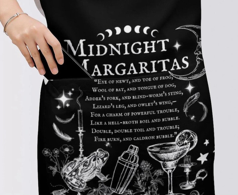May include: A black apron featuring white graphic illustrations of a moon, stars, a frog, a cocktail shaker, and a margarita glass. The text reads Midnight Margaritas followed by a poem starting with Eye of newt, and toe of frog, wool of bat.