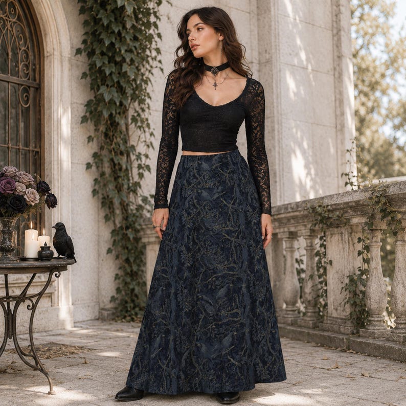 May include: A black long-sleeved lace crop top with a V-neckline paired with a floor-length dark blue maxi skirt featuring a detailed tonal pattern of branches and birds. The outfit is completed with a black choker necklace and black shoes.