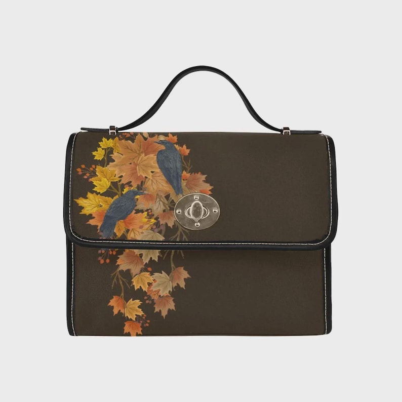 May include: A dark brown satchel with a top handle and a silver-toned metal turn-lock closure. The front flap features a printed design of two black birds perched among a cluster of vibrant orange, yellow, and brown autumn maple leaves.