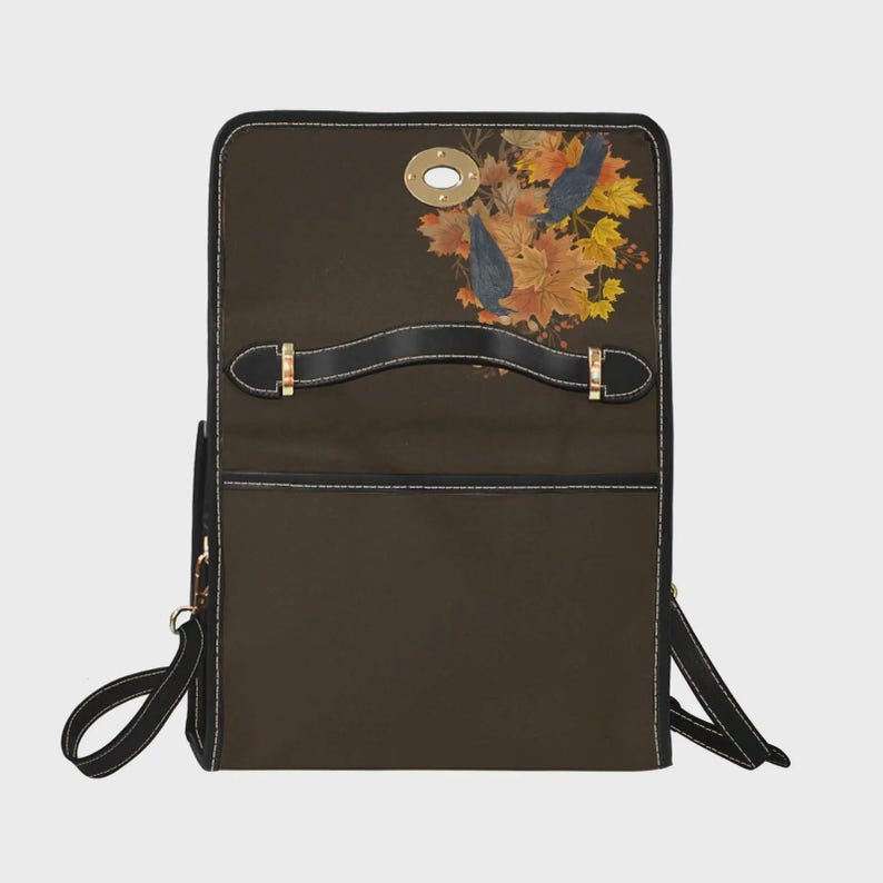 May include: A dark brown backpack featuring a top handle and a flap closure with a gold-toned metal turn-lock. The flap is decorated with a printed graphic of two dark birds perched among a cluster of autumn-colored maple leaves in shades of orange.