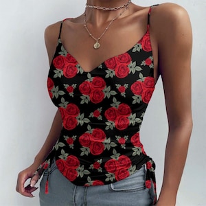 May include: Black camisole top with a red rose pattern. The top has a V-neck and thin straps. The fabric is decorated with a repeating pattern of red roses and green leaves. The sides of the top have adjustable drawstring details.