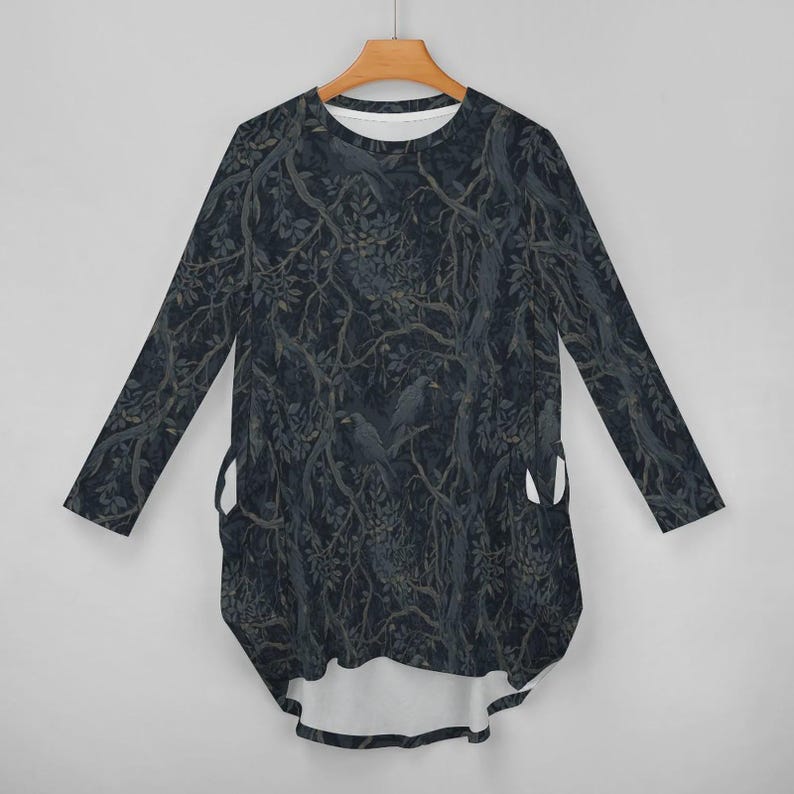 May include: A long-sleeve tunic top featuring a dark, moody forest pattern with black crows perched on tangled tree branches. The garment has a rounded neckline, a high-low hemline, and two side pockets integrated into the dark, leafy print design.