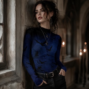 May include: A long-sleeved mock neck top in a deep blue hue featuring a repeating black bird silhouette pattern. The garment is paired with black denim jeans secured by a black leather belt with a silver buckle, and accessorized with a silver necklace.