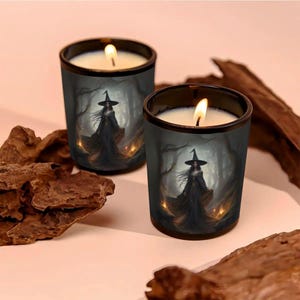 May include: Two lit candles in black glass holders. Each candle features a witch illustration in a dark forest setting. The candles have a white wax and a flickering flame, creating a spooky atmosphere.