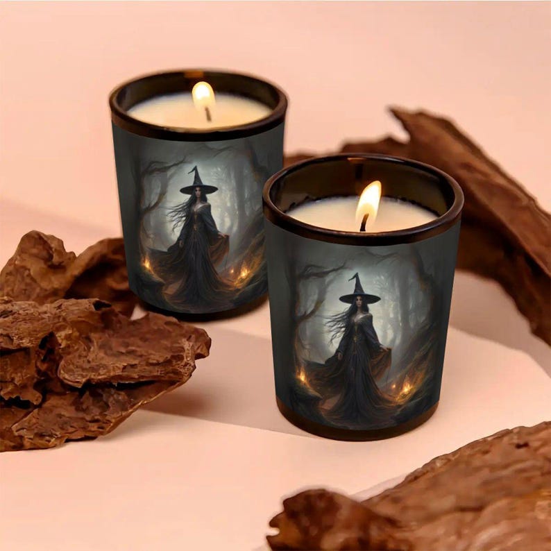 May include: Two lit candles in black glass holders. Each candle features a witch illustration in a dark forest setting. The candles have a white wax and a flickering flame, creating a spooky atmosphere.