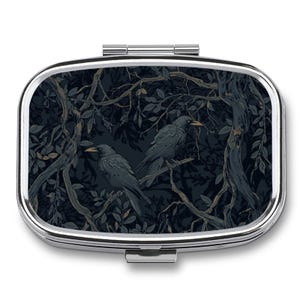 May include: A silver pill box featuring a detailed illustration of two crows perched on a branch within a forest setting. The artwork uses a palette of dark blues and greens.