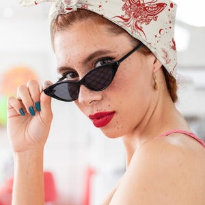 May include: A cream-colored headscarf featuring a red graphic print of a moth, crescent moons, and stars. The fabric is tied at the top of the head in a knot. The design includes intricate line work and celestial motifs across the entire surface.