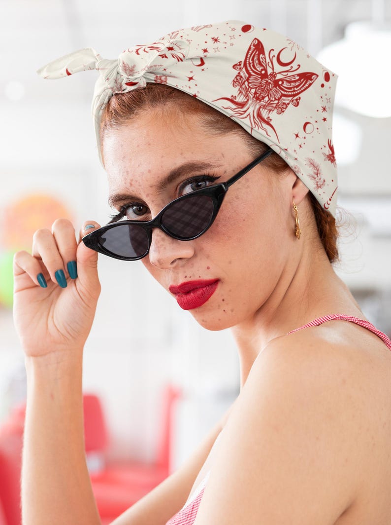 May include: A cream-colored headscarf featuring a red graphic print of a moth, crescent moons, and stars. The fabric is tied at the top of the head in a knot. The design includes intricate line work and celestial motifs across the entire surface.