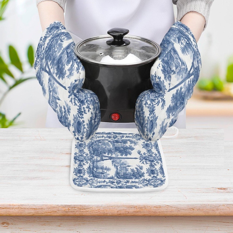 May include: A pair of blue and white oven mitts and a matching pot holder with a toile pattern. The mitts are holding a black pot with a glass lid. The pot holder is on a white surface.