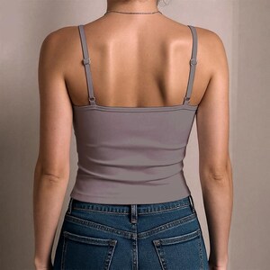 May include: A solid mauve-coloured camisole top featuring thin, adjustable shoulder straps with silver-toned metal sliders. The garment is worn tucked into a pair of dark blue denim jeans with visible back pockets and a classic stitched waistband.