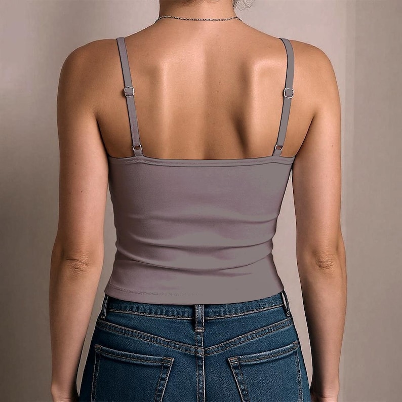 May include: A solid mauve-coloured camisole top featuring thin, adjustable shoulder straps with silver-toned metal sliders. The garment is worn tucked into a pair of dark blue denim jeans with visible back pockets and a classic stitched waistband.
