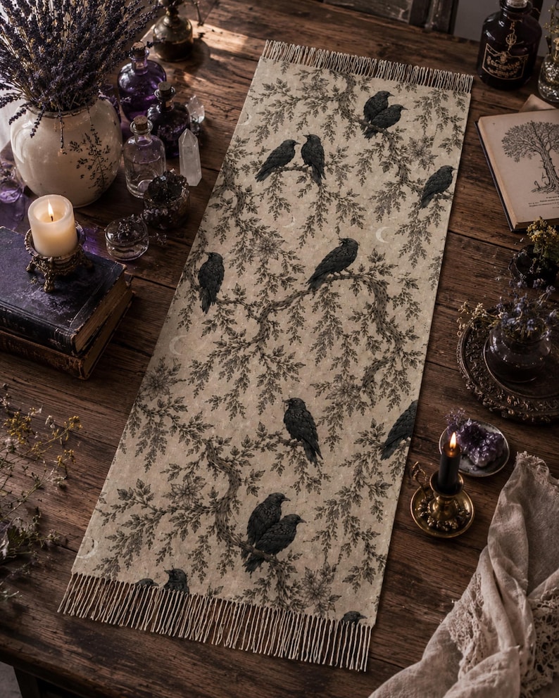Op de afbeelding: Een rechthoekige tafelloper met een beige achtergrond en een herhalende zwarte print van kraaien die op boomtakken zitten. De loper heeft aan beide korte zijden franjes en toont een gedetailleerd botanisch patroon met vogels.