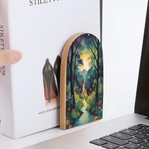May include: A wooden arched bookmark featuring a watercolor painting of a forest scene with a castle, a full moon, and a winding path. The bookmark is placed between the pages of a book with the text "STILETTOS" and "AN ILLUSTRATED HI".
