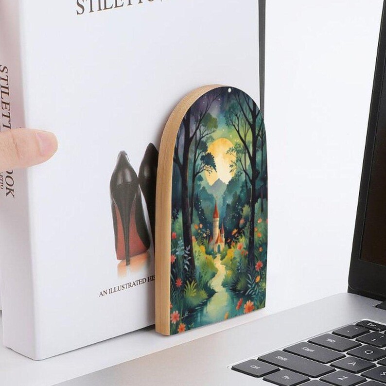May include: A wooden arched bookmark featuring a watercolor painting of a forest scene with a castle, a full moon, and a winding path. The bookmark is placed between the pages of a book with the text "STILETTOS" and "AN ILLUSTRATED HI".
