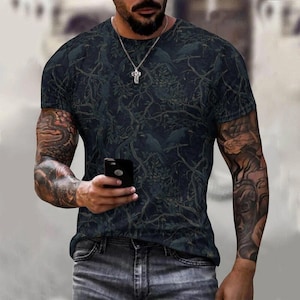 Pode incluir: Uma t-shirt de manga curta com gola redonda, apresentando um padr&atilde;o escuro e intrincado de aves em silhueta pousadas em ramos de &aacute;rvores densos e entrela&ccedil;ados. A t-shirt &eacute; combinada com cal&ccedil;as de ganga cinzentas e um colar de prata.