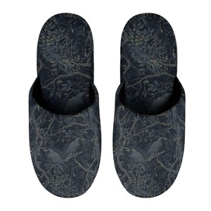 May include: A pair of slip-on slippers featuring a dark navy blue and charcoal gray pattern of crows perched on tree branches with dense foliage. The design covers the entire exterior of the footwear, showcasing a detailed botanical and avian motif.
