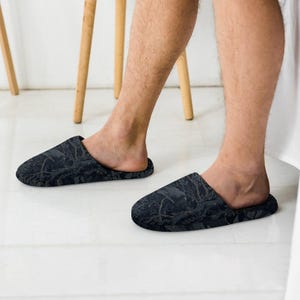 May include: A pair of slip-on slippers featuring a dark charcoal gray and black botanical pattern with intricate illustrations of leaves and branches. The slippers have a rounded toe design and a low-profile silhouette, providing full coverage.