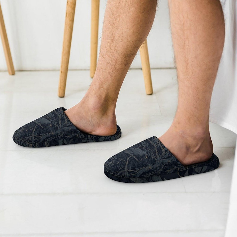 May include: A pair of slip-on slippers featuring a dark charcoal gray and black botanical pattern with intricate illustrations of leaves and branches. The slippers have a rounded toe design and a low-profile silhouette, providing full coverage.