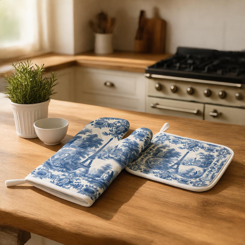 May include: A blue and white oven mitt and a square pot holder featuring a toile de jouy pattern with illustrations of the Eiffel Tower and floral motifs. Both items have a white fabric trim and a small loop for hanging on a wooden surface.