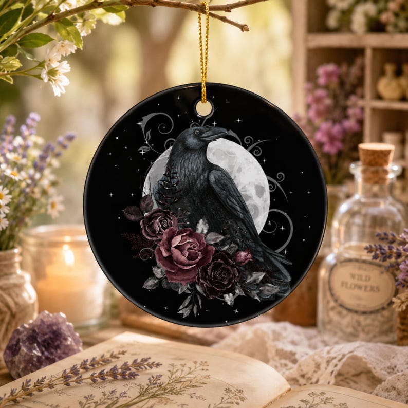 May include: A round black ceramic ornament featuring a detailed illustration of a black raven perched among dark red roses against a large white full moon background. The ornament is suspended by a thin gold cord and includes decorative swirls.
