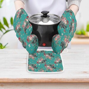 May include: A pair of teal oven mitts and a matching square pot holder featuring a floral pattern with red roses and dark bird illustrations. The mitts are shown holding a black electric pot with a glass lid and a visible red power switch.