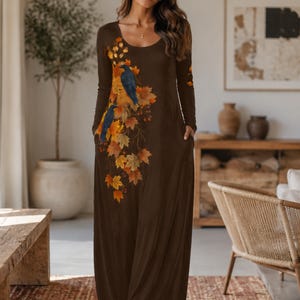 Könnte beinhalten: Ein bodenlanges, braunes Maxikleid mit langen Ärmeln, Rundhalsausschnitt und Seitentaschen. Die Vorderseite des Kleides ist mit einem Druck von zwei dunkelblauen Vögeln verziert, die zwischen orangefarbenen, gelben und roten Herbstblättern sitzen.