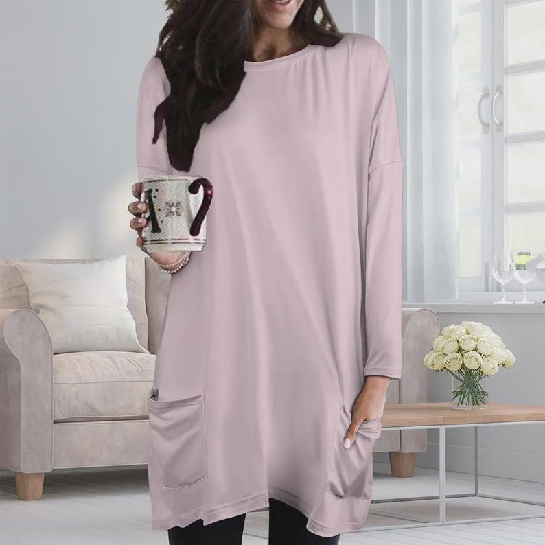 May include: A long-sleeved, light pink tunic top featuring a round neckline and two large patch pockets on the front. The garment has a loose, relaxed fit with dropped shoulders and a straight hemline that falls below the hip area.