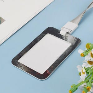Puede incluir: Un porta credencial vertical rectangular con un patr&oacute;n abstracto y jaspeado en tonos negros y grises. El soporte incluye una pieza de pl&aacute;stico blanco en la parte superior conectada a una correa delgada de color claro para colgar.