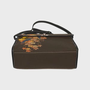 May include: A dark brown rectangular shoulder bag featuring a printed design of autumn maple leaves in shades of orange, yellow, and red, along with a small blue bird. The bag has black trim, white stitching, and a metallic turn-lock closure.
