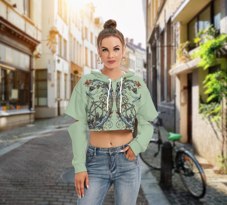 May include: A light green cropped hoodie featuring long sleeves with cut-out shoulders and a white drawstring. The front displays a detailed graphic print of mushrooms, swirling vines, and dragonfly illustrations in muted earth tones and blue accents.