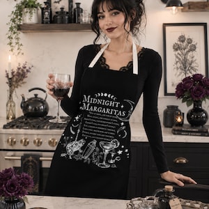 May include: A black apron featuring white graphic illustrations of a frog, a cocktail shaker, and a margarita glass. The text reads Midnight Margaritas along with a potion recipe. The apron has white neck straps and a central printed design.