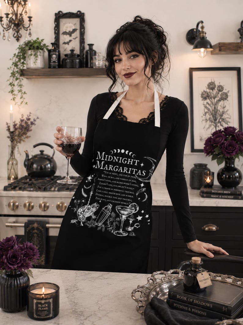 May include: A black apron featuring white graphic illustrations of a frog, a cocktail shaker, and a margarita glass. The text reads Midnight Margaritas along with a potion recipe. The apron has white neck straps and a central printed design.