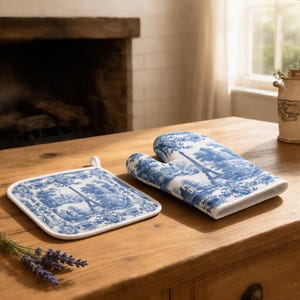 May include: A square pot holder and an oven mitt featuring a blue and white toile de Jouy pattern. The fabric design depicts a landscape with the Eiffel Tower, trees, and buildings. Both items have white fabric trim and a small hanging loop.