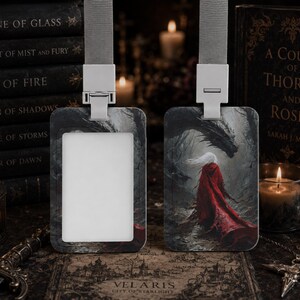Puede incluir: Dos portatarjetas rectangulares con cordones grises. Uno tiene un inserto rectangular blanco, mientras que el otro muestra una ilustraci&oacute;n de fantas&iacute;a de una figura con una capa roja larga frente a un gran drag&oacute;n oscuro en un bosque.