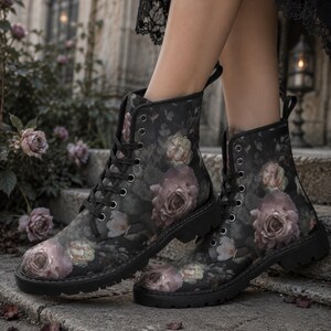 May include: A pair of black ankle boots featuring a dark floral pattern with muted pink and white roses. The boots have black laces, silver-toned metal eyelets, a pull tab at the heel, and a thick, black lug sole with a deep tread pattern.