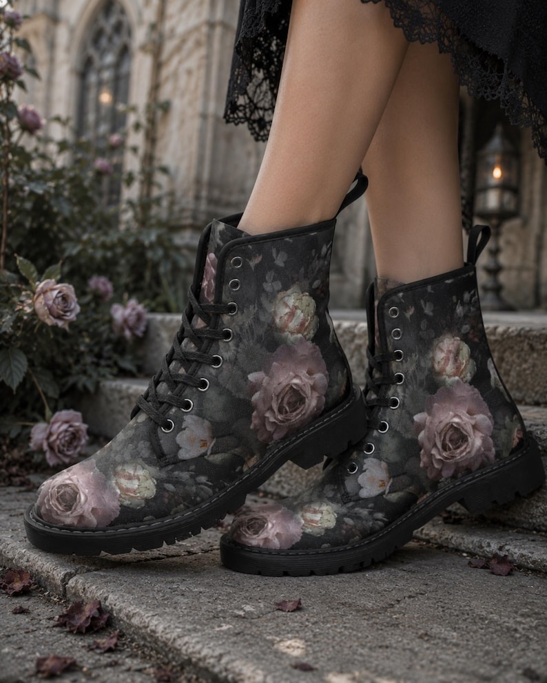 May include: A pair of black ankle boots featuring a dark floral pattern with muted pink and white roses. The boots have black laces, silver-toned metal eyelets, a pull tab at the heel, and a thick, black lug sole with a deep tread pattern.