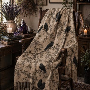Op de afbeelding: Een beige plaid gedrapeerd over een houten stoel, met een herhalend patroon van zwarte kraaien zittend op donkere, bladerrijke takken. De deken heeft een textuur met franjes aan de onderkant en kleine motieven van een halve maan.