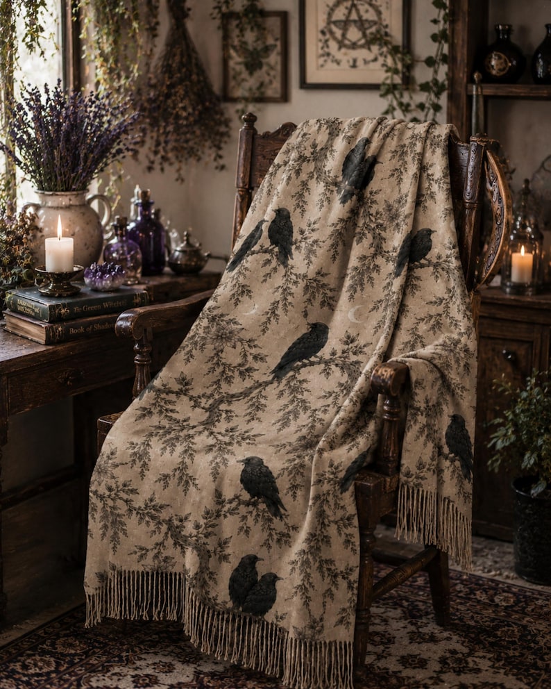 Op de afbeelding: Een beige plaid gedrapeerd over een houten stoel, met een herhalend patroon van zwarte kraaien zittend op donkere, bladerrijke takken. De deken heeft een textuur met franjes aan de onderkant en kleine motieven van een halve maan.