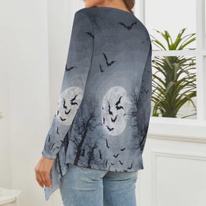 May include: A long-sleeve top featuring a gray and black gradient print of a night sky with two full moons, silhouettes of bare tree branches, and numerous flying bats. The garment has a rounded neckline and a slightly asymmetrical hemline.
